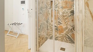 Een hotelkamer met een houten visgraatvloer, een douchecabine met glazen wanden en bloemenbehang, en een witte deur.