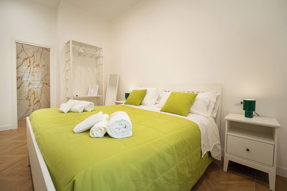 Ein ordentlich bezogenes Bett mit grüner Bettwäsche, ein weißer Nachttisch mit grünem Becherhalter und eine Wand mit Marmoroptik.