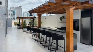 Een dakterras met een lange bar, zwarte barkrukken, een moderne keukenhoek en een stadszicht op de achtergrond.