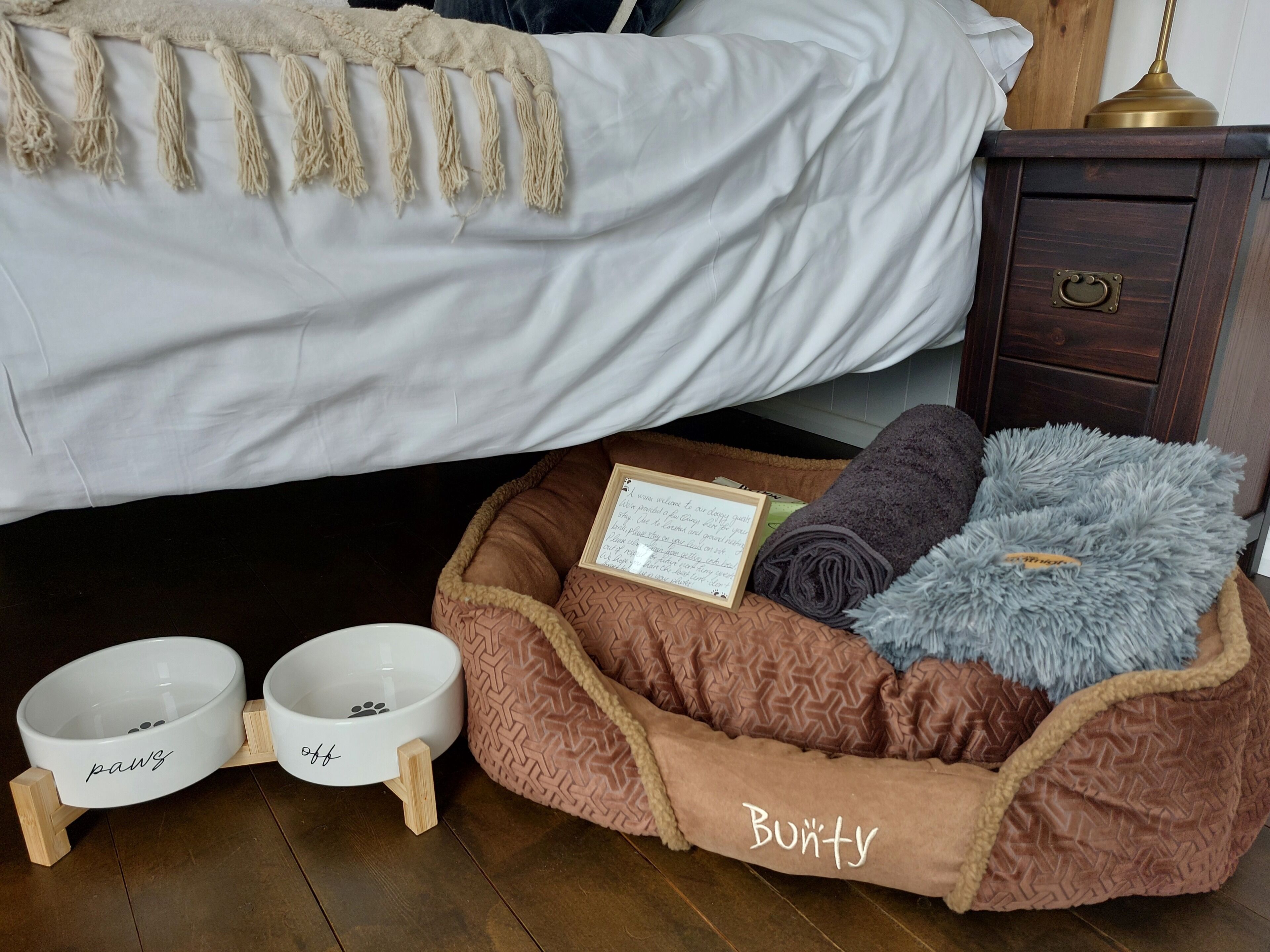 A pet bed with a name tag, two pet bowls, a wooden nightstand, and a bed with a white sheet and a beige throw blanket.