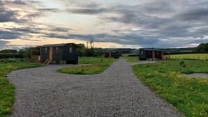 A gravel path leading to a row of small wooden cabins with a grassy field and cloudy sky in the background. - Captain's Cabin Shepherd's Hut with Hot Tub, on the edge of the North York Moors (Pinchinthorpe)