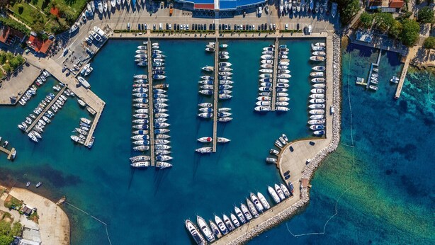 Eine Luftaufnahme einer Marina mit zahlreichen vertäuten Booten, einer Mole und einem Gebäude mit blauem Dach.