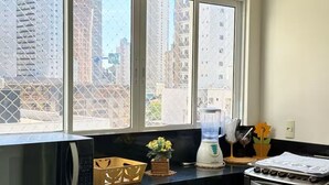 Eine Küche mit Mikrowelle, Mixer und Herd, ein Fenster mit Blick auf hohe Gebäude und eine kleine Pflanze auf der Arbeitsfläche.