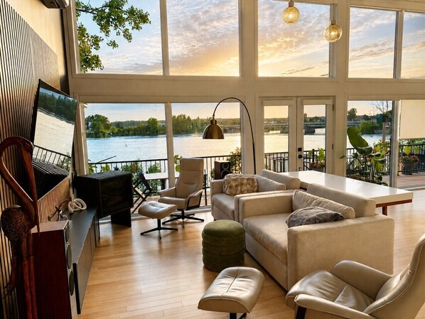 A spacious living area with a large window overlooking a body of water, a fireplace, and a television.