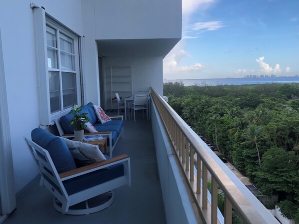 A balcony with a sofa, chair, and dining table, overlooking a dense green landscape and a distant city skyline. - Recently redecorated 2 bedroom Apt with Stunning Views & Prime Location (Key Biscayne)
