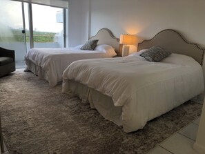 Ein Hotelzimmer mit zwei Betten, einem Nachttisch mit Lampe, Blick auf die Stadt durch ein großes Fenster und einem Teppichboden.
