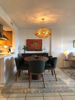 A dining area with a round table, four chairs, a chandelier, a painting, and a kitchen in the background. - Recently redecorated 2 bedroom Apt with Stunning Views & Prime Location (Key Biscayne)