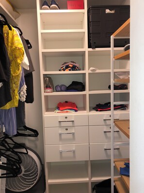 Ein weißer Stauraum mit mehreren Regalen und Schubladen, der gefaltete Kleidung, einen Ventilator und eine rote Kiste enthält.