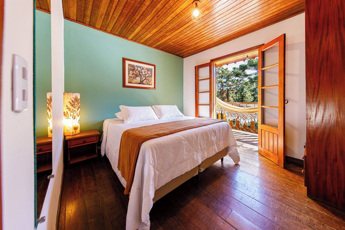 Un dormitorio con techo de madera, cama con ropa blanca, mesita de noche con lámpara y vista a un jardín a través de una puerta.