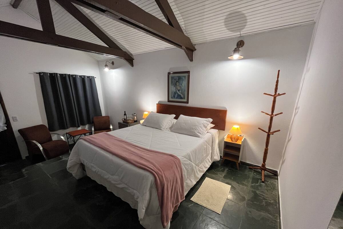 Un dormitorio con cama, una silla, una mesita de noche, una lámpara y un perchero.