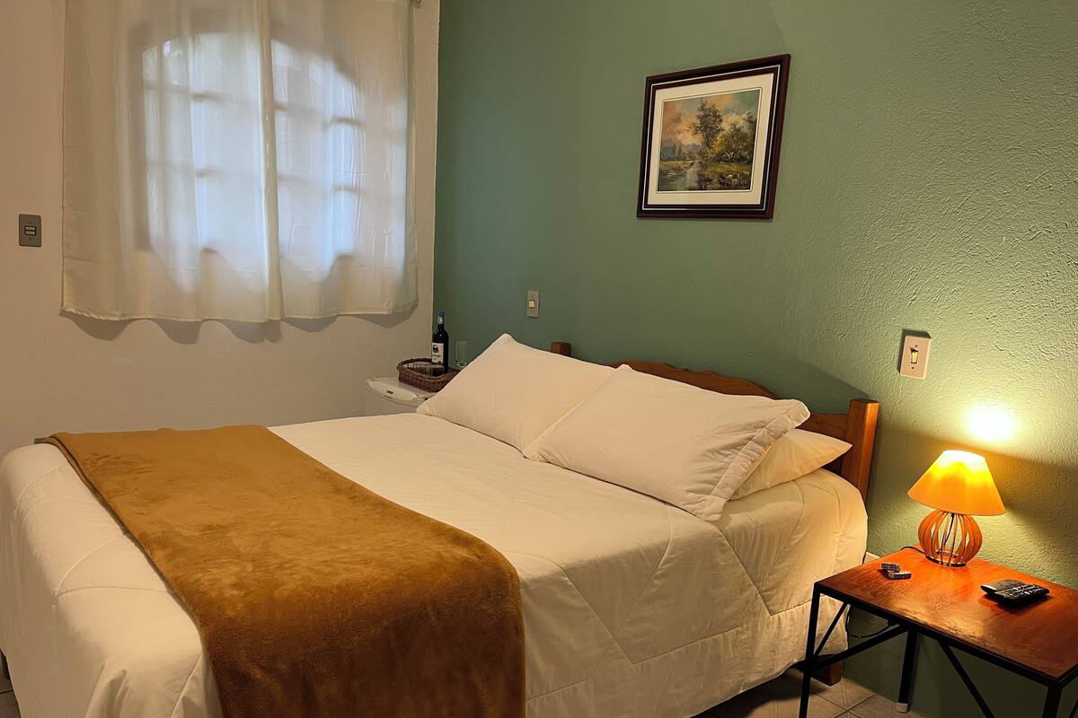 Una cama bien hecha con una manta marrón, una mesita de noche con una lámpara y un cuadro en la pared.