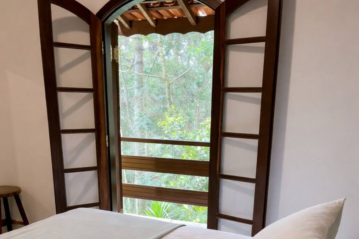 Una cama con sábanas y almohadas blancas, una ventana con marco de madera y vista a la vegetación exterior.