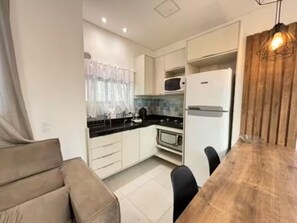 A compact kitchen with white cabinets, a microwave, a refrigerator, a dining table with chairs, and a wooden accent wall. - Fully furnished apartment in Meia Praia – Easy access and garage. (Itapema)