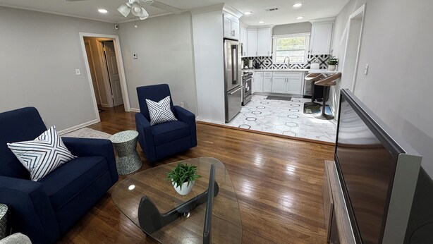 Een moderne woonkamer met een bank, fauteuil en salontafel, een keuken met witte kasten en een betegelde spatwand, en een plafondventilator.