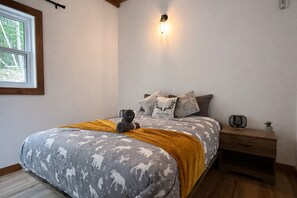 A bedroom with a bed featuring a nature-themed bedspread, a bedside table with a lamp, and a window with a view of trees.