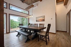 A dining area with a large table, chairs, and a bench. There are windows allowing natural light and a view of the outdoors.
