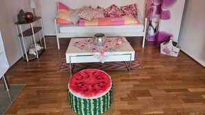 Een gezellige kamer met een witte bank, een ronde tafel met een tafelkleed met bloemen, een watermeloen krukje, en een klein plankje met decoratieve objecten.