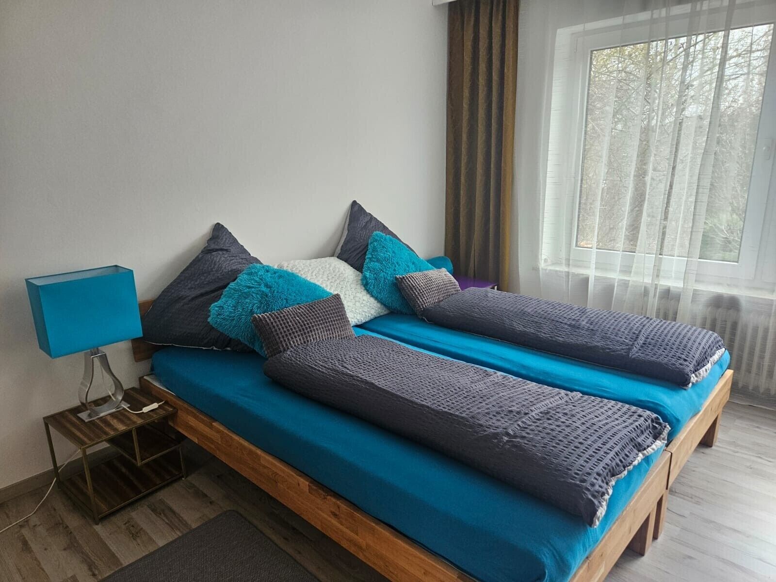 Ein Doppelbett mit blauer Bettdecke und mehreren Kissen, ein Nachttisch mit Lampe sowie ein Fenster mit Vorhängen.