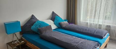 Ein Doppelbett mit blauer Bettdecke und mehreren Kissen, ein Nachttisch mit Lampe sowie ein Fenster mit Vorhängen.