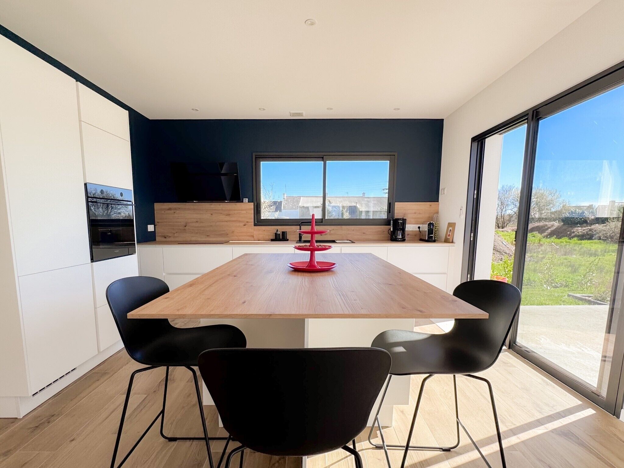 A modern kitchen with a wooden table, black chairs, and a large window offering a view of the outdoors.