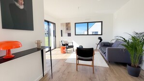 A modern living room with a sofa, a chair, a table with a red object, and a large window. - Ty Nevez – Holiday Home with Private Terrace, Garden & Wi-Fi in Penmarch (Penmarch)