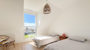 A bedroom with two beds, a wicker chair, a bedside table with a red lamp, and a large window with a view of a body of water. - Ty Nevez – Holiday Home with Private Terrace, Garden & Wi-Fi in Penmarch (Penmarch)