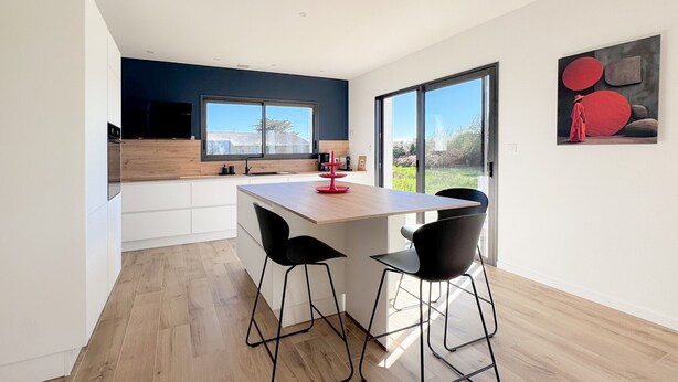 A modern kitchen with a wooden island, black bar stools, and a large window. - Ty Nevez – Holiday Home with Private Terrace, Garden & Wi-Fi in Penmarch (Penmarch)