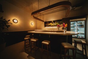 A bar area with stools, a counter, a refrigerator stocked with bottles, and a wall-mounted shelf with bottles.