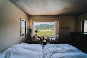 A room with a bed, two chairs, a table, and a view of a green landscape through large windows.