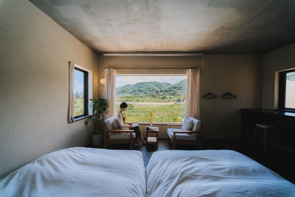 A room with a bed, two chairs, a table, and a view of a green landscape through large windows. - Semilla NAGO OKINAWA (Nago)