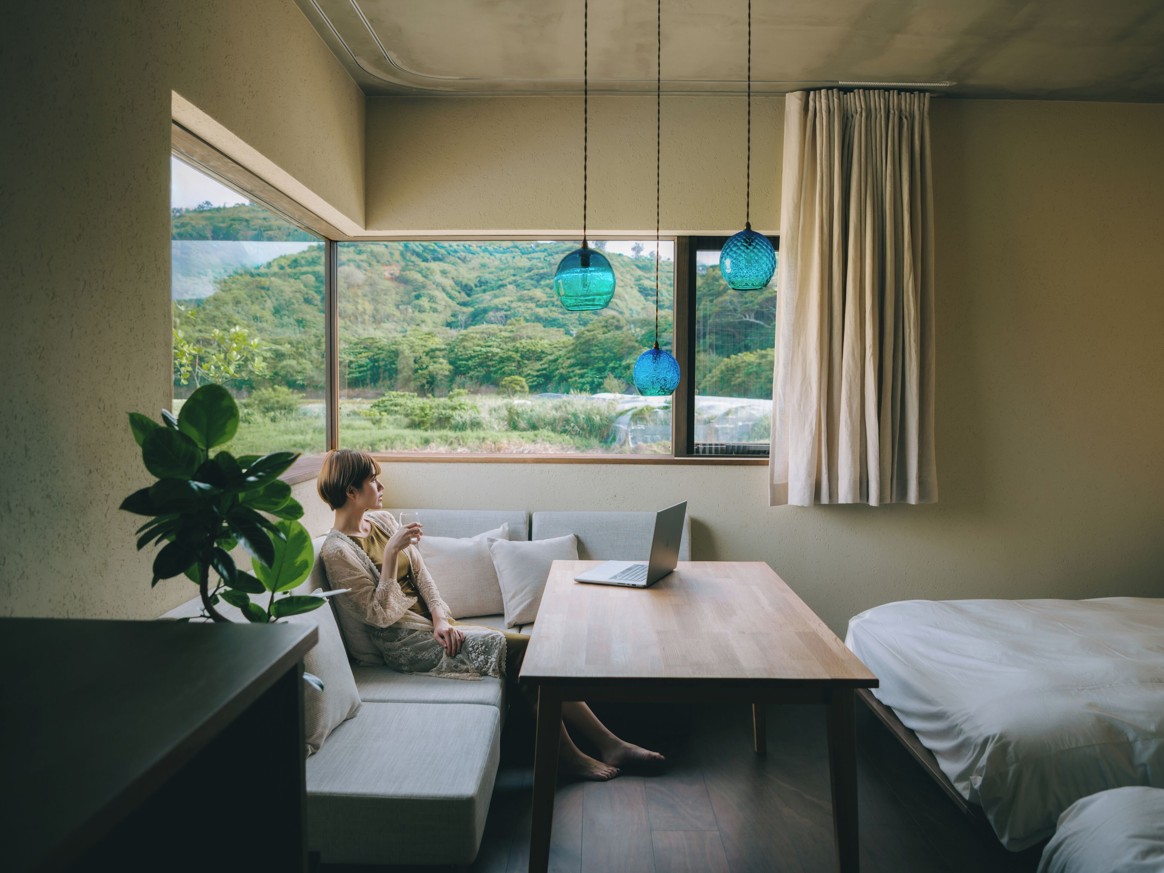 A room with a view of a green landscape, a sofa, a table with a laptop, and a hanging light fixture.