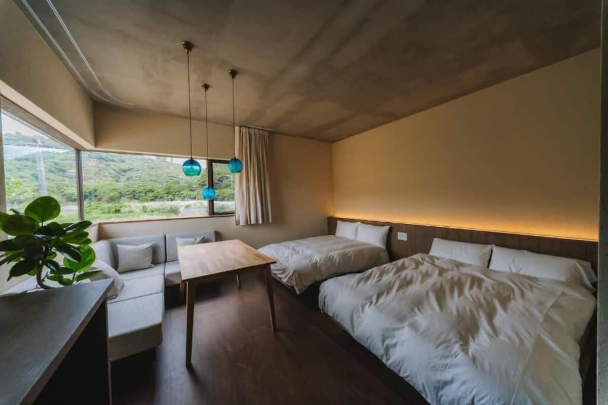 A modern bedroom with a sofa, two beds, a table, and a view of greenery outside.