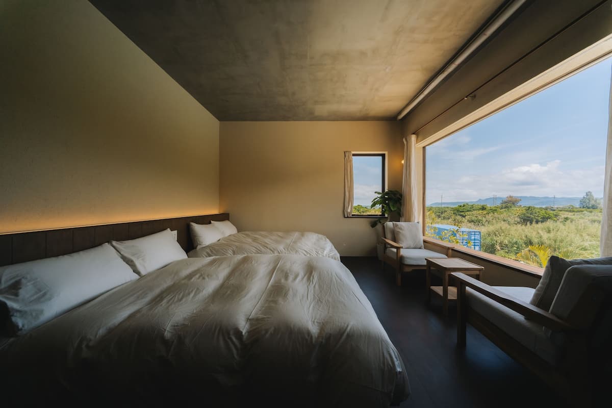 A bedroom with a large bed, a desk, a chair, and a window with a view of greenery.