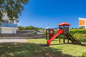 A playground with a red slide and a wooden structure, surrounded by grass and trees. - Sea Breeze Funtana Studio Apartment A10 (Funtana)