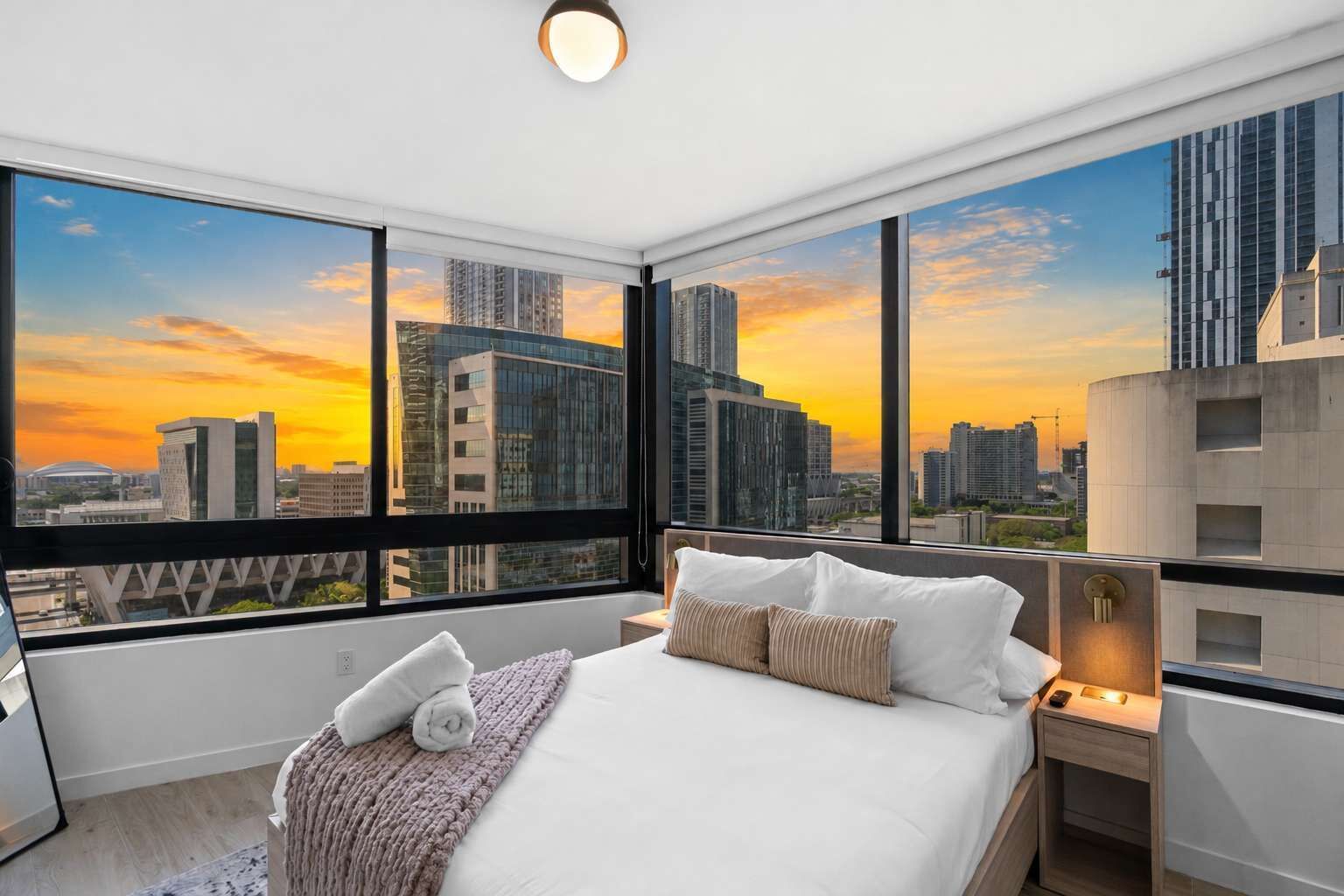 Habitación de hotel moderna con una cama grande, una mesita de noche y vistas a un horizonte urbano al atardecer.