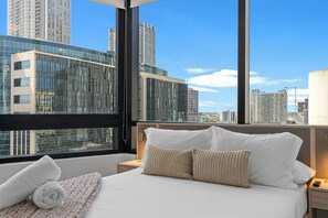 A hotel room with a large bed, a view of tall buildings, and a window with a black frame. - 1-Bed | Metromover & Brightline Access (Miami)