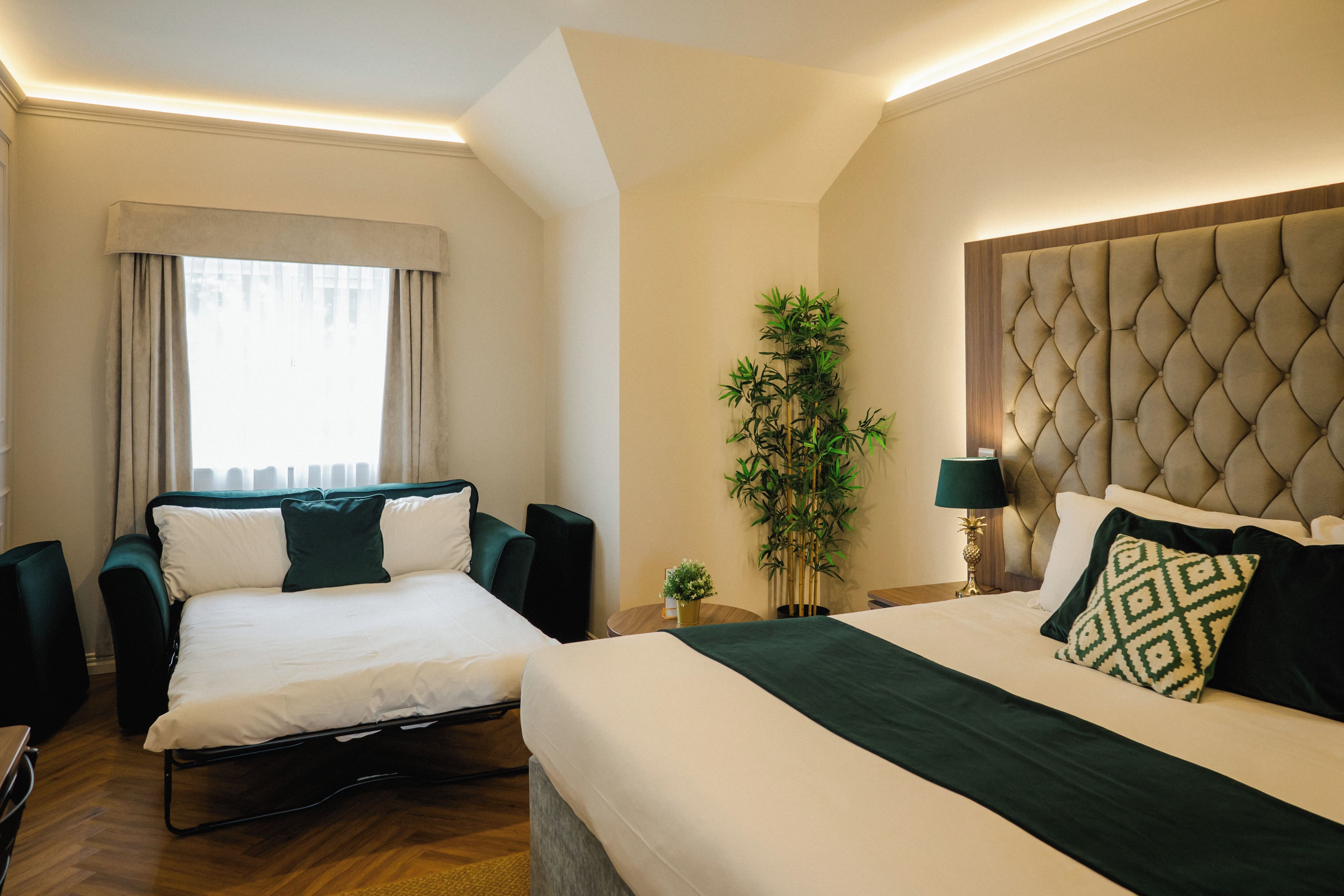 A hotel room with two beds, a large window with curtains, a potted plant, and a nightstand with a lamp.