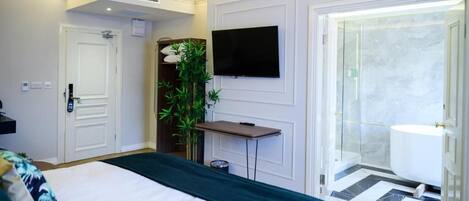 Habitación de hotel con cama, televisor en la pared, zona de ducha y una mesita con una planta.