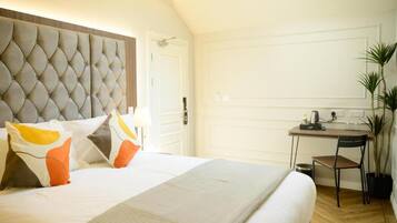 A hotel room with a tufted headboard, a bed with white bedding and orange pillows, a desk with a chair, and a potted plant.