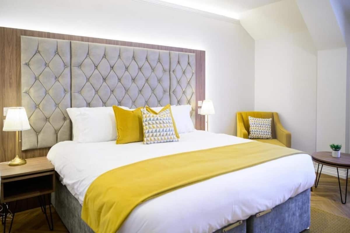 Ein Hotelzimmer mit einem großen Bett, einem gepolsterten Kopfteil, zwei Nachttischen mit Lampen, einem kleinen runden Tisch mit Pflanze und einem gelben Sessel.