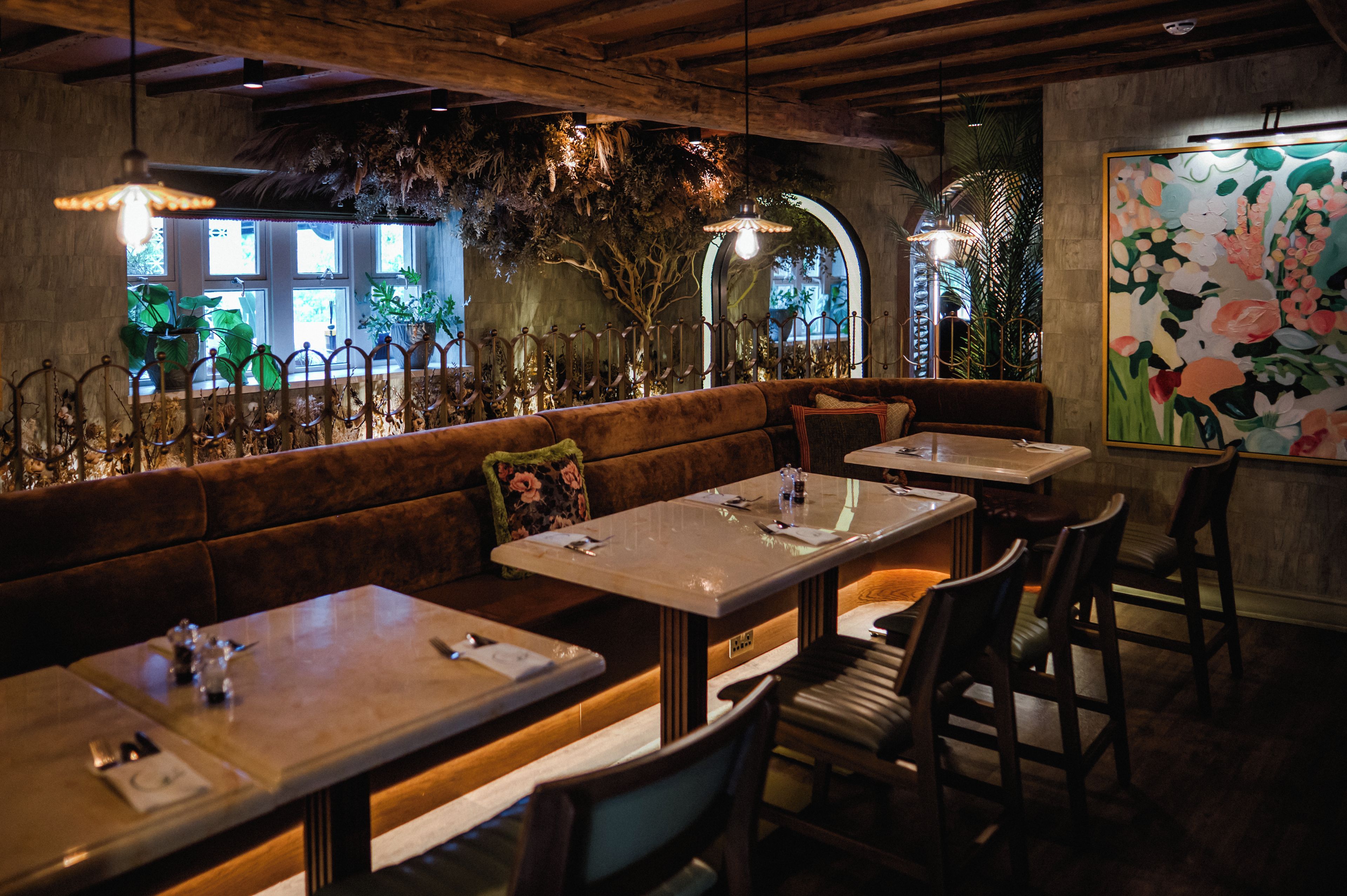 A restaurant interior with wooden beams, a bench seating area, tables, chairs, and a large floral painting on the wall.