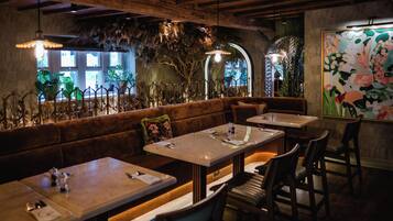 A restaurant interior with wooden beams, a bench seating area, tables, chairs, and a large floral painting on the wall.