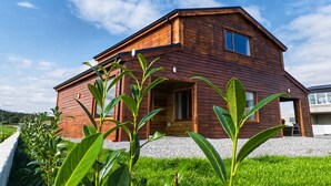 A two-story wooden house with a sloped roof, a front porch, and a small garden area with green plants.