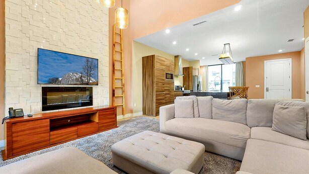 A modern living room with a flat-screen TV, a wooden entertainment unit, a beige sofa, and a kitchen in the background. - Spacious Luxury 4BR Suite Near Disney (Kissimmee)