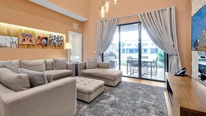 A modern living room with a sectional sofa, a flat-screen TV, and a large window with curtains. - Spacious Luxury 4BR Suite Near Disney (Kissimmee)