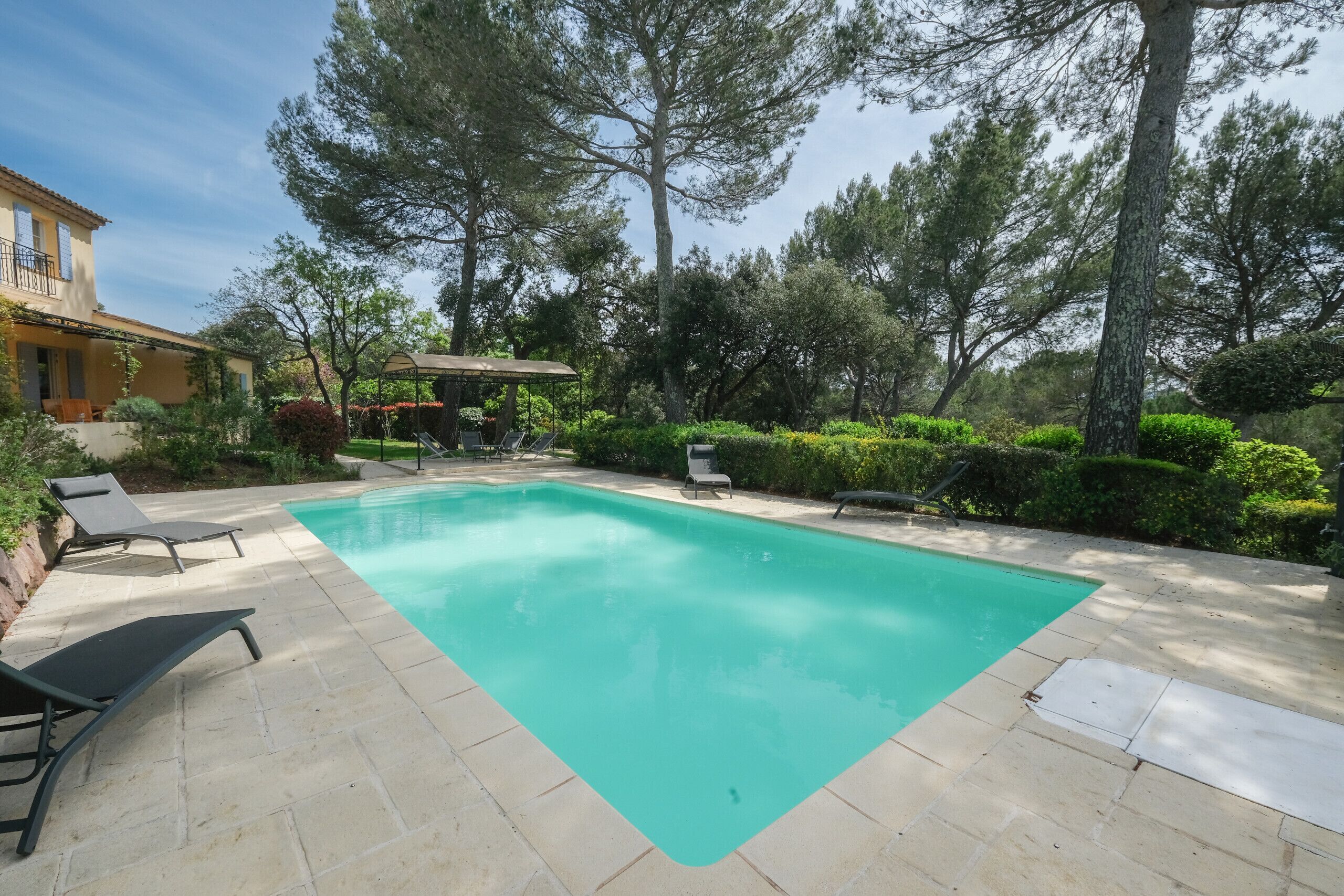 Une piscine entourée d’arbres et d’un bâtiment, avec des chaises longues à proximité.