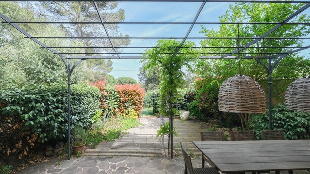 Een terras buiten met stenen vloer, een houten tafel en een pergola.