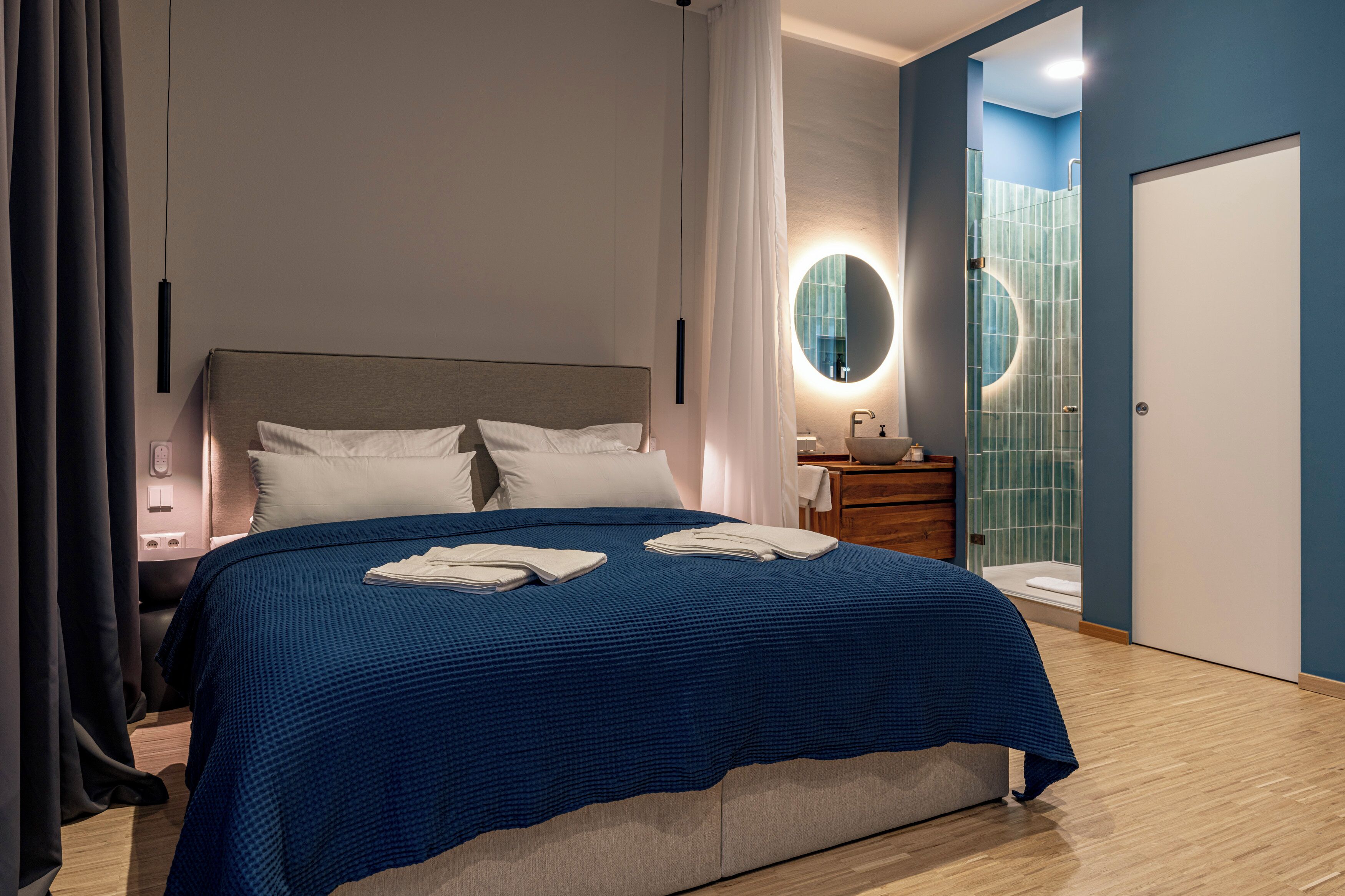 Ein modernes Hotelzimmer mit einem großen Bett, einem Nachttisch, einem Spiegel, einem Badezimmer mit einer blauen Duschkabine und einer Tür.