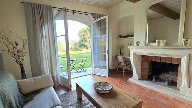 Ein Zimmer mit Kamin, Esstisch, Sofa, Stuhl und Blick auf eine grüne Landschaft durch eine geöffnete Tür.