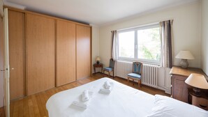 Una habitación de hotel con una cama grande, un armario de madera, dos sillas, una mesa con una lámpara y una ventana con cortinas.